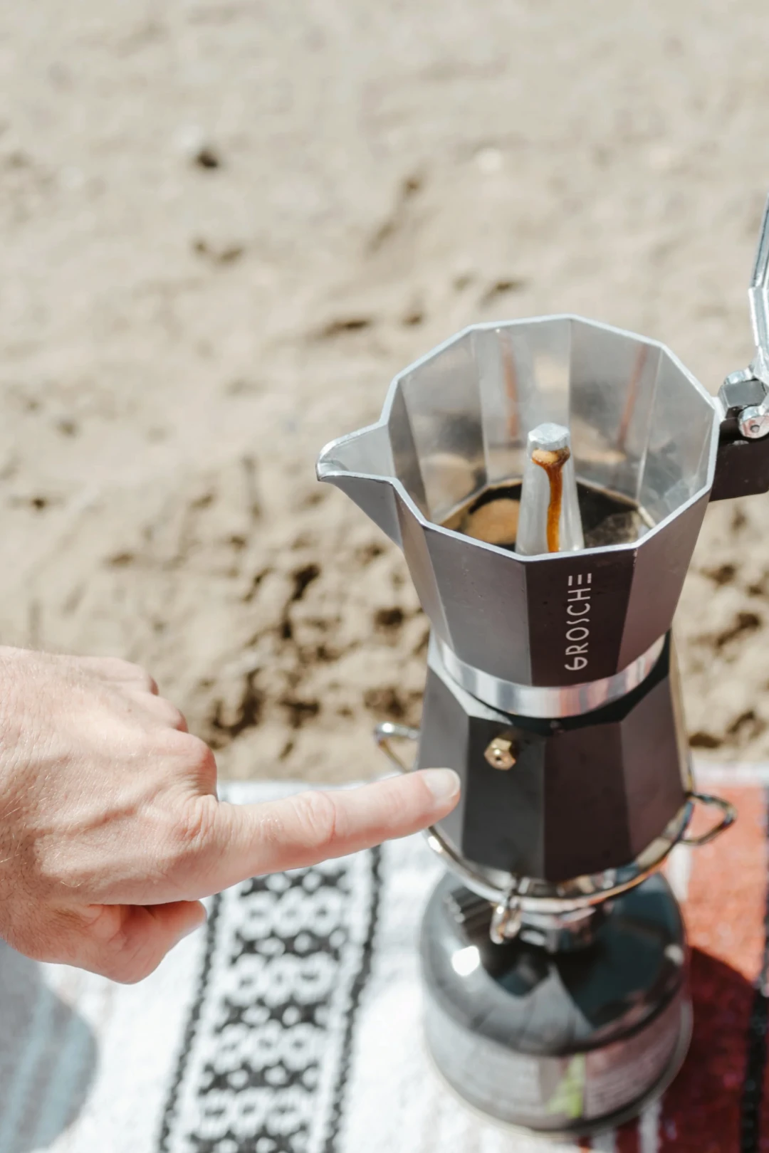 A close-up of a silver aluminum moka pot with a hand pointing at it, set on a patterned blanket with a sandy beach background.