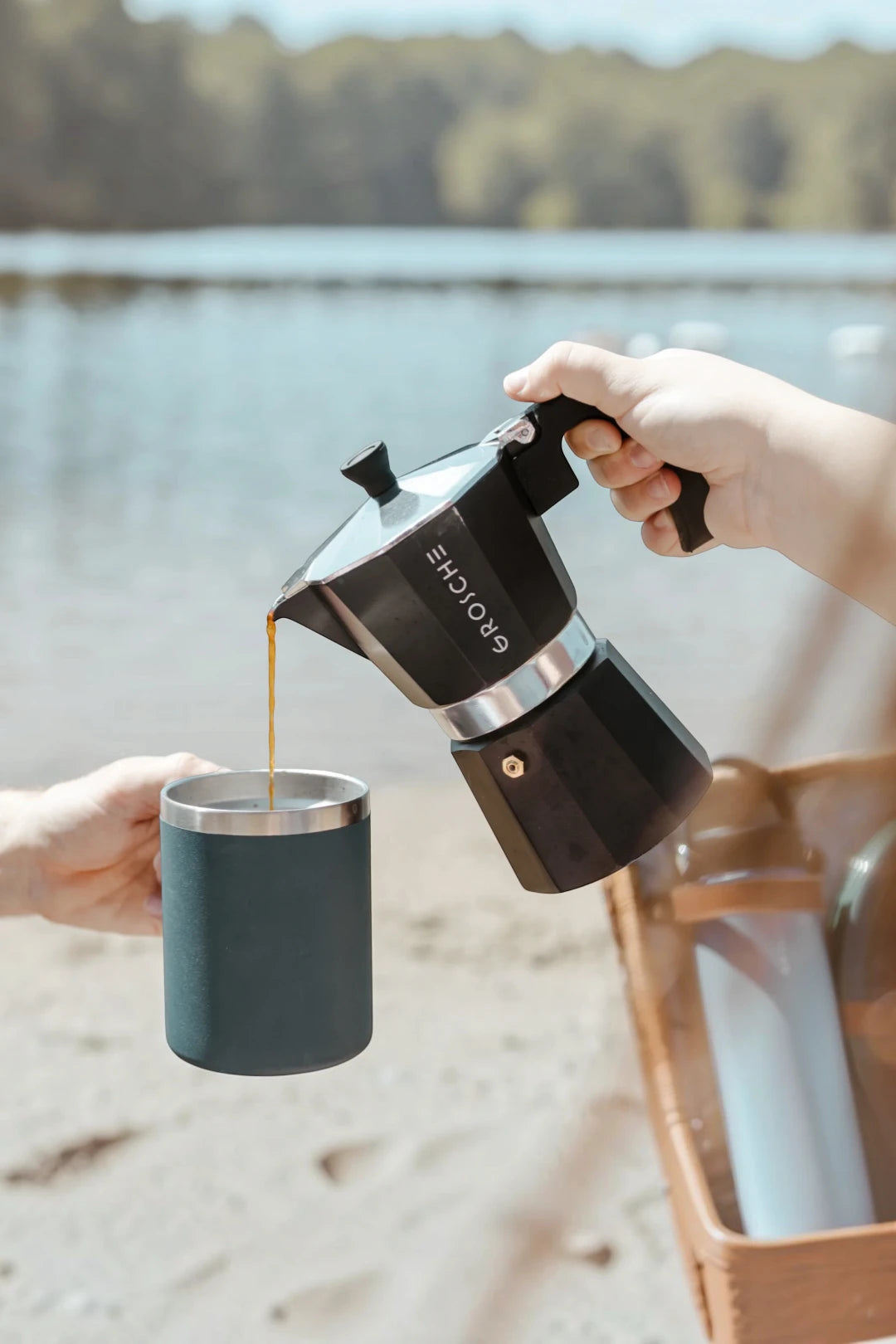 A person pouring coffee from a black and silver moka pot into a dark teal insulated mug, with a blurred outdoor lakeside background.