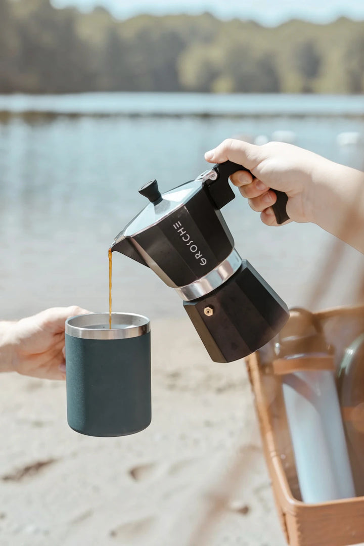 A person pouring coffee from a black and silver moka pot into a dark teal insulated mug, with a blurred outdoor lakeside background.