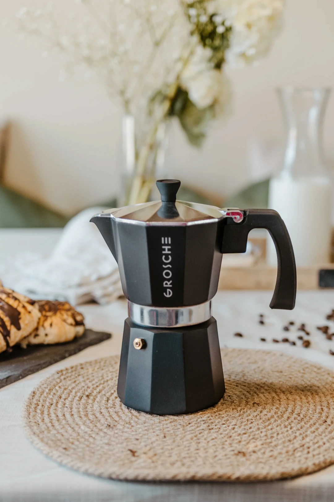 A black GROSCHE moka pot coffee maker sits on a round woven placemat on a table, with blurred coffee beans, a glass of milk, and flowers in the background.