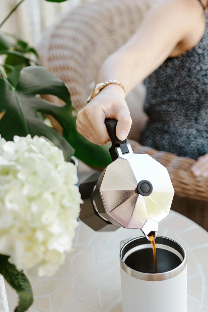A person pouring hot coffee from an octagonal silver moka pot into a white insulated travel mug on a table, with green leaves and white flowers in the foreground.