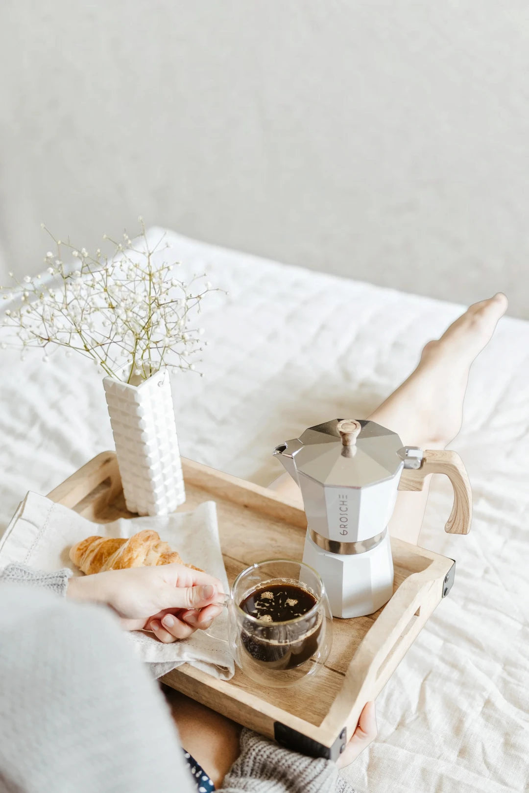 A white moka pot and a glass of coffee on a wooden tray, styled on a bed with a person's hands and feet visible, alongside a croissant and a vase of baby's breath.