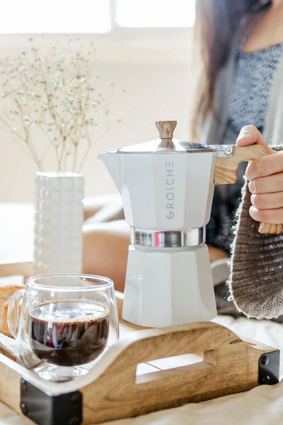 A white moka pot with a wooden print handle and wooden print top knob sits on a wooden tray next to a glass of coffee, with a person holding the handle in a softly lit indoor setting.