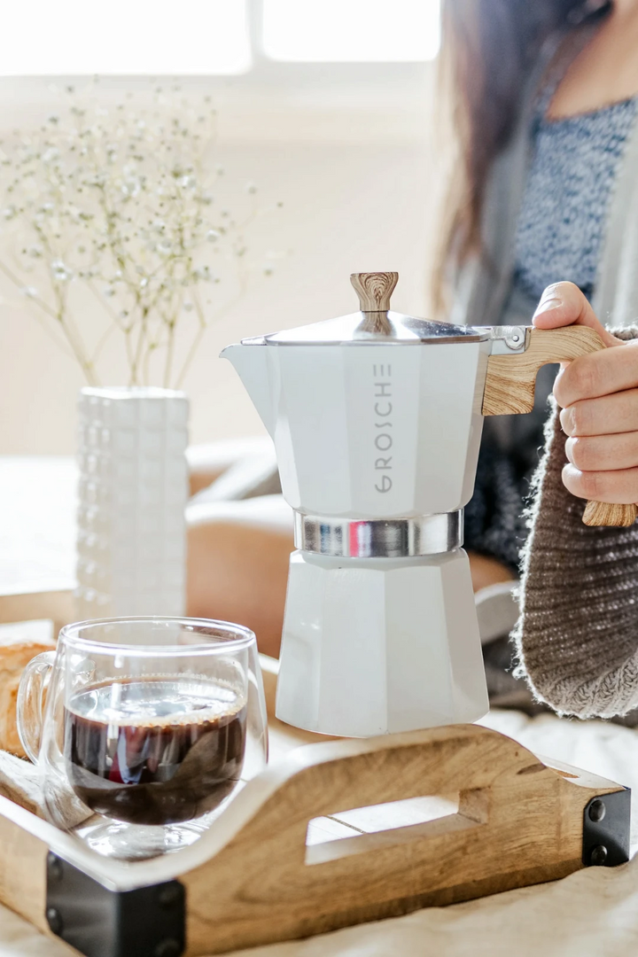 A white moka pot with a wooden print handle and wooden print top knob sits on a wooden tray next to a glass of coffee, with a person holding the handle in a softly lit indoor setting.