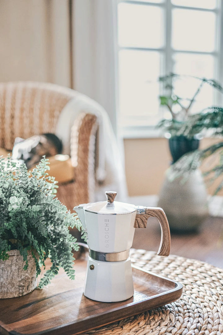 A white stovetop moka pot with a light wood print handle sits on a wooden tray on a woven table in a softly lit living room. A potted fern is in the foreground, and a wicker chair and window are blurred in the background.