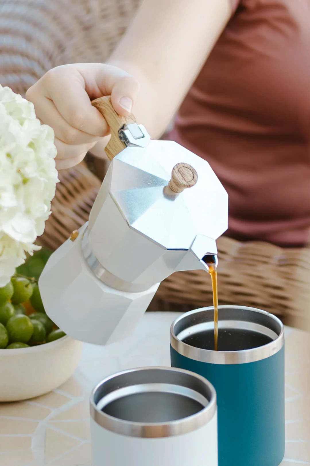 A hand pouring coffee from a silver moka pot into two insulated tumblers, one teal and one white, on a table with a bowl of green grapes and white flowers in the background.