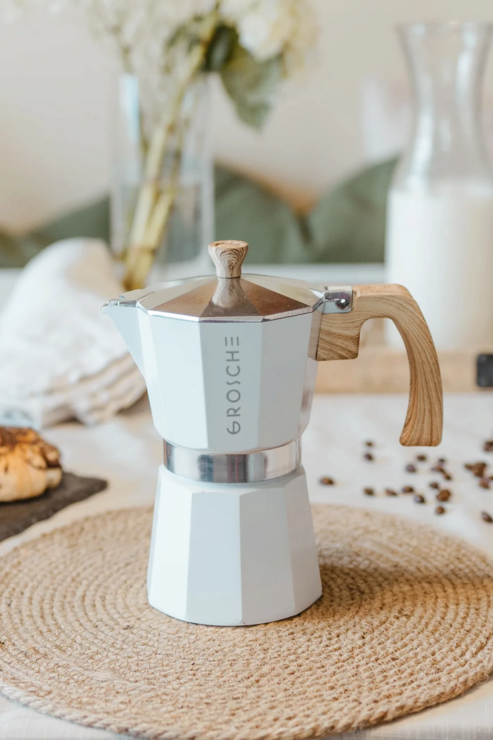A white and stainless steel moka pot with a light wood handle and lid knob sits on a woven placemat in a softly styled kitchen setting.