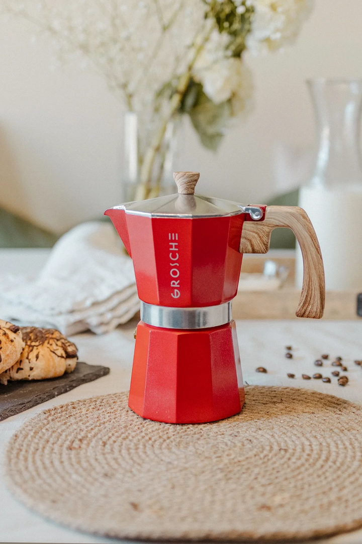 A red and silver stovetop moka pot with a wooden handle and lid knob sits on a woven placemat on a table, with coffee beans scattered nearby and a blurred background showing flowers, bread, and a glass of milk.