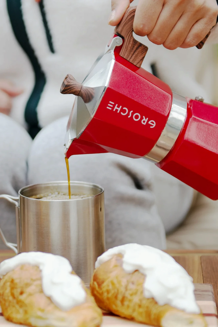 A red and silver moka pot with a wooden handle is being used by a person to pour coffee into a stainless steel mug. Pastries topped with white cream are visible in the foreground on a wooden surface.