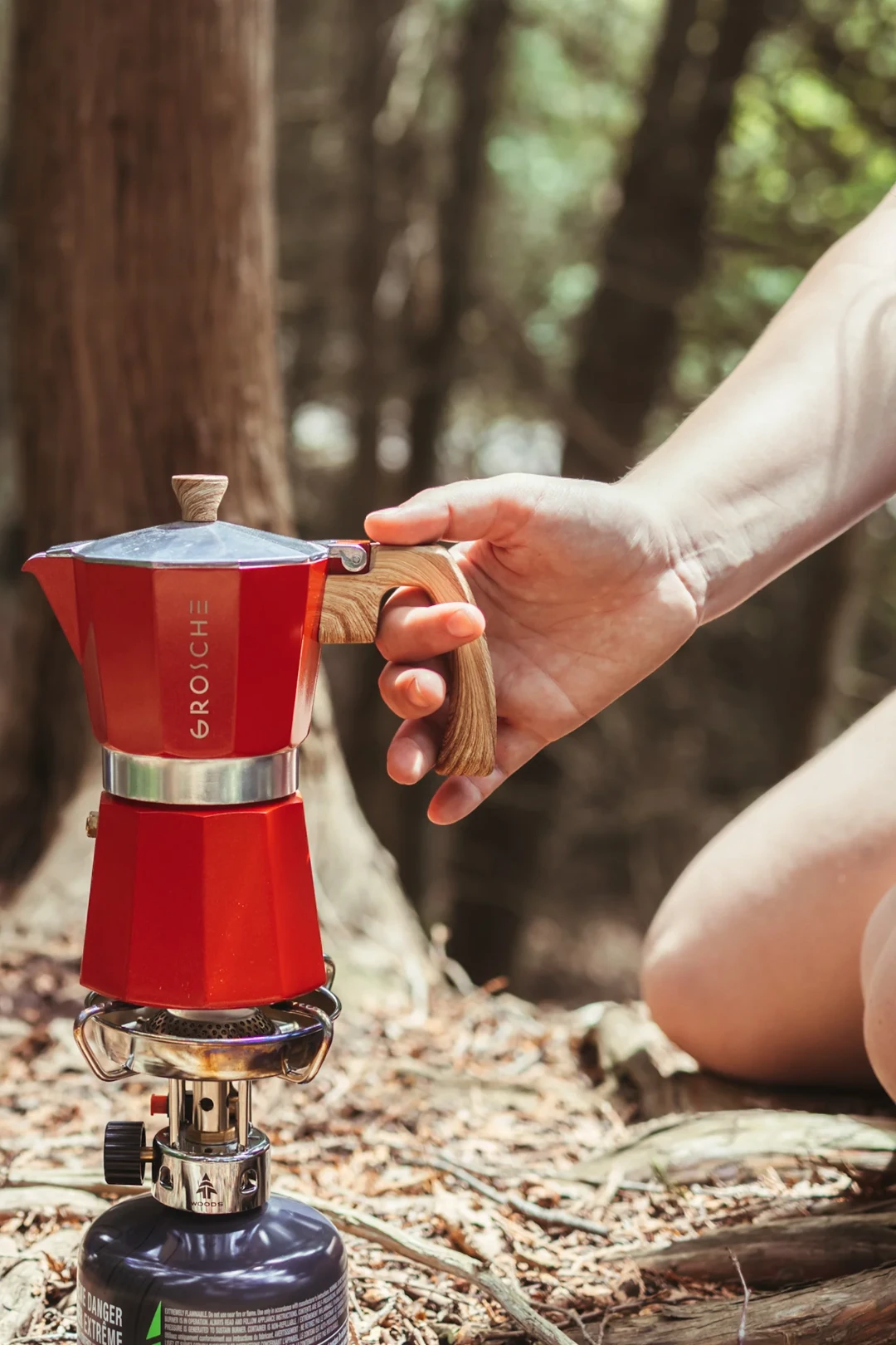 A red and silver moka pot with a wooden handle sits on a camping stove, with a person's hand holding the handle in a forest setting.