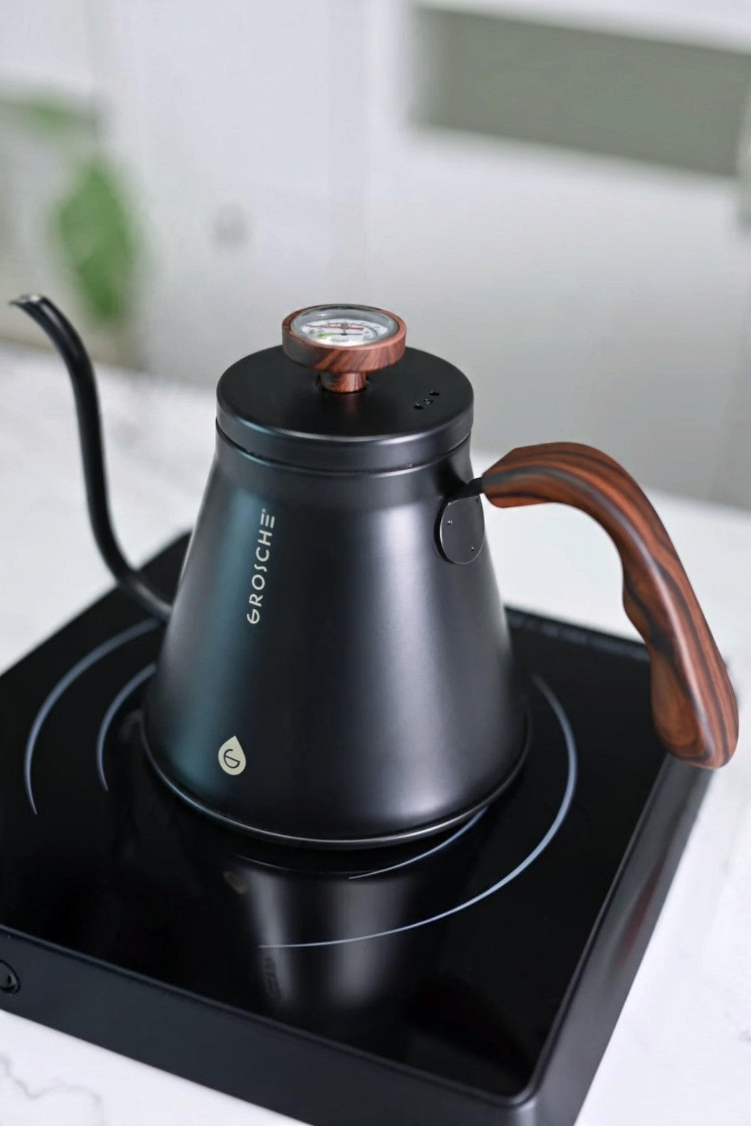 A matte black gooseneck kettle with a copper-toned temperature gauge and a brown wooden handle (Thermoplastic elastomer (TPE)), sitting on a black induction cooktop. The brand name 'GROSCHE' is printed vertically on the side of the kettle.