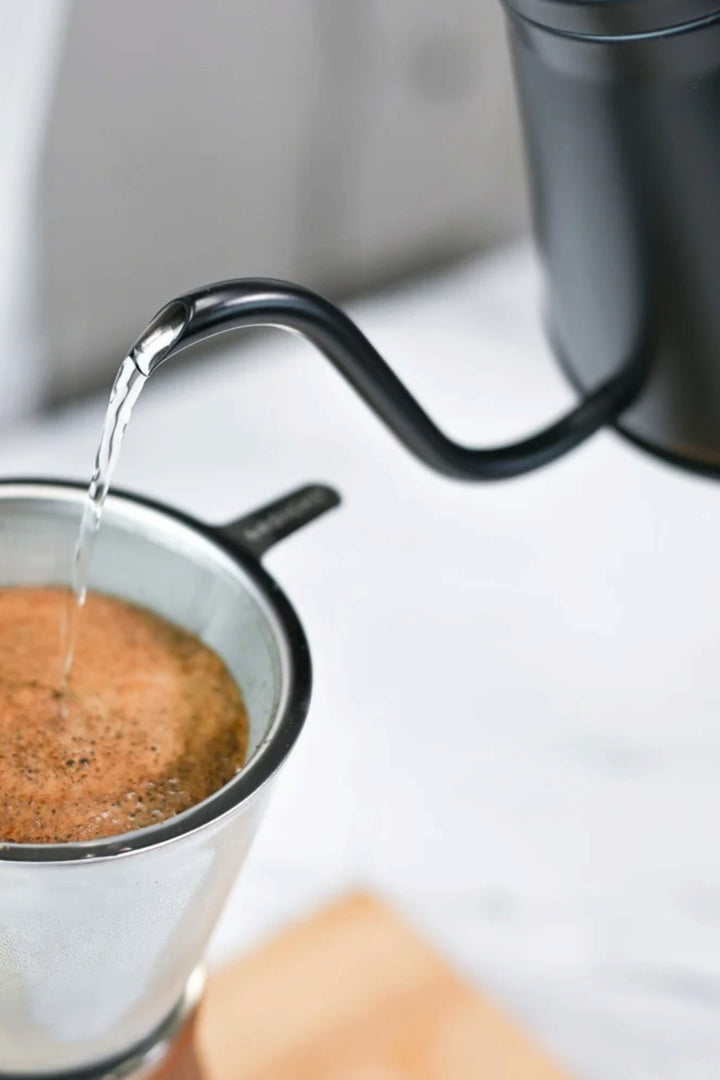 A close-up of water being poured from a black gooseneck kettle into a metal pour-over filter containing coffee grounds, on a light marble-like surface.