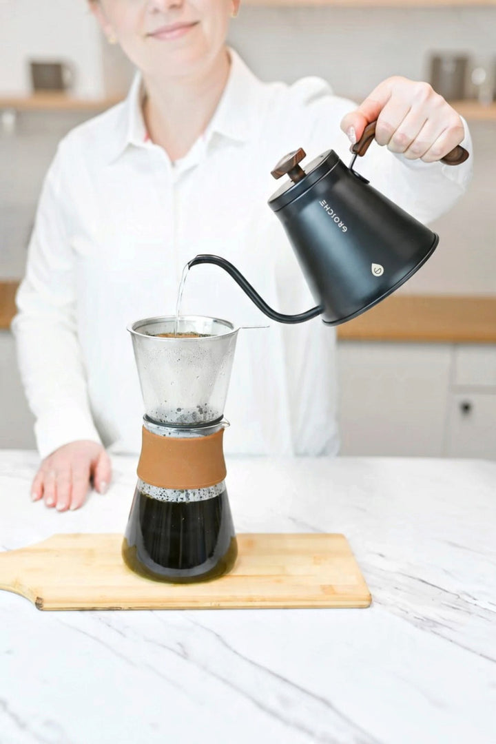 A person in a white shirt pours hot water from a matte black gooseneck kettle into a pour-over coffee dripper set on a wooden board. The kettle has the brand name 'GROSCHE' visible on its side.