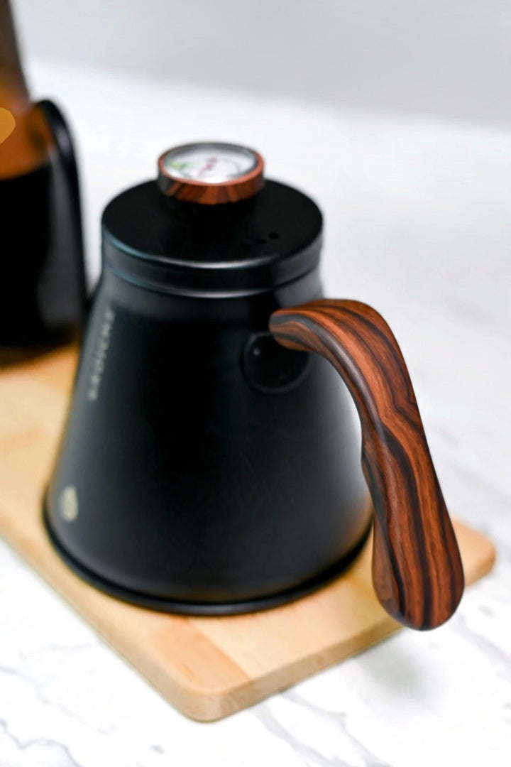 A matte black coffee kettle with a distinctive dark brown wood grain handle and lid knob, resting on a light wooden coaster.
