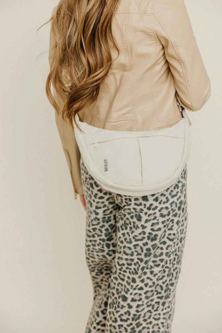 A woman shown from the back wearing a light beige faux-leather jacket, leopard-print pants, and a cream-colored crescent-shaped crossbody bag with a small black logo.